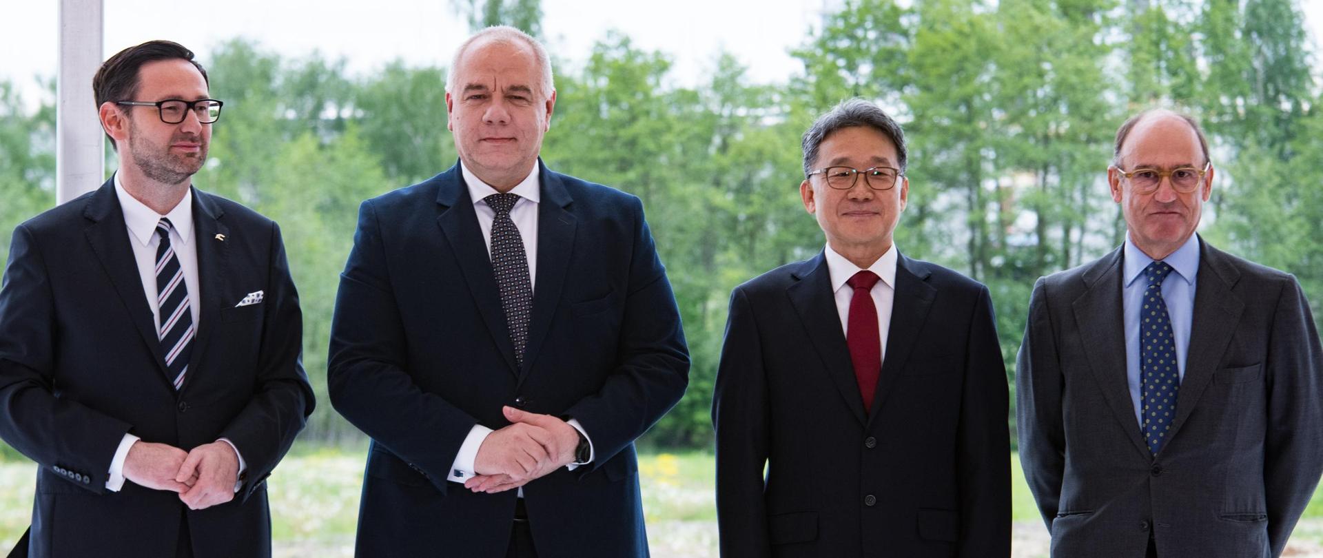 Uczestnicy konferencji pozują do wspólnego zdjęcia. Czterech mężczyzn w garniturach stoi za stołem, na którym rozłożone są dokumenty. Na blacie stoją też trzy pudełka z piórami.