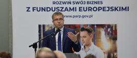 Dyrektor Departamentu Programów Wsparcia Innowacji i Rozwoju w MFiPR Grzegorz Orawiec stoi przy mównicy i przemawia do mikrofonu, za jego plecami baner informujący o rozwoju biznesu z Funduszami Europejskimi
