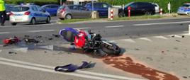 Na zdjęciu przód leżącego na prawym boku rozbitego motocykla. Widoczny czerwony bak paliwa i fioletowe fragmenty osłon. Na jezdni brunatne ślady rozsypanego sorbentu. W tle na sąsiednim pasie ruchu jadące samochody osobowe. Po lewej stronie zdjęcia widoczny tył i prawy bok pojazdu policji i fragment pleców policjanta. Po prawej stronie zdjęcia tył kolejnego policyjnego samochodu.