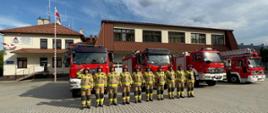 Na zdjęciu widoczni strażacy na tle budynku komendy i samochodów pożarniczych. Po lewej stronie zdjęcia zdjęcia masz z flagą na środku salutujący strażak dowódca zmiany służbowej obok niego po prawej stronie zmiana służbowa w jednym szeregu. Strażacy ubrani w czerwone hełmy i ubrania specjalne koloru piaskowego.