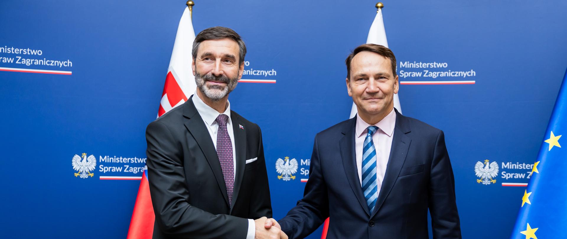 Na zdjęciu są ministrowie spraw zagranicznych Polski i Słowacji. Są ubrani w garnitury. W tle jest granatowa ścianka z napisem Ministerstwo Spraw Zagranicznych