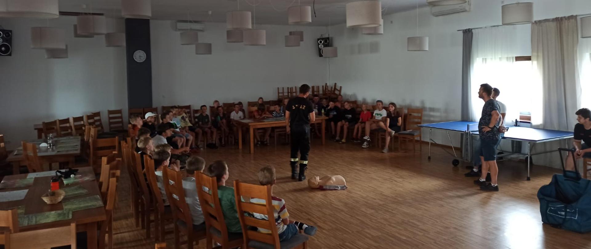 Zdjęcie przedstawia strażaka oraz dzieci i ich opiekunów. Znajdują się oni w dużym pomieszczeniu, gdzie są krzesełka i stoliki. Dzieci słuchają strażaka, który do nich mówi. Strażak ubrany jest w ubranie koszarowe. Obok strażaka znajduje się manekin do udzielania pierwszej pomocy.