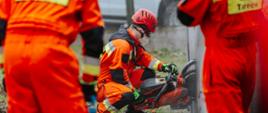 Strażacy ubrani w pomarańczowe kombinezony na głowie mają ubrane czerwone kaski, przy użyci przecinarki spalinowej wykonują otwór w płycie betonowej