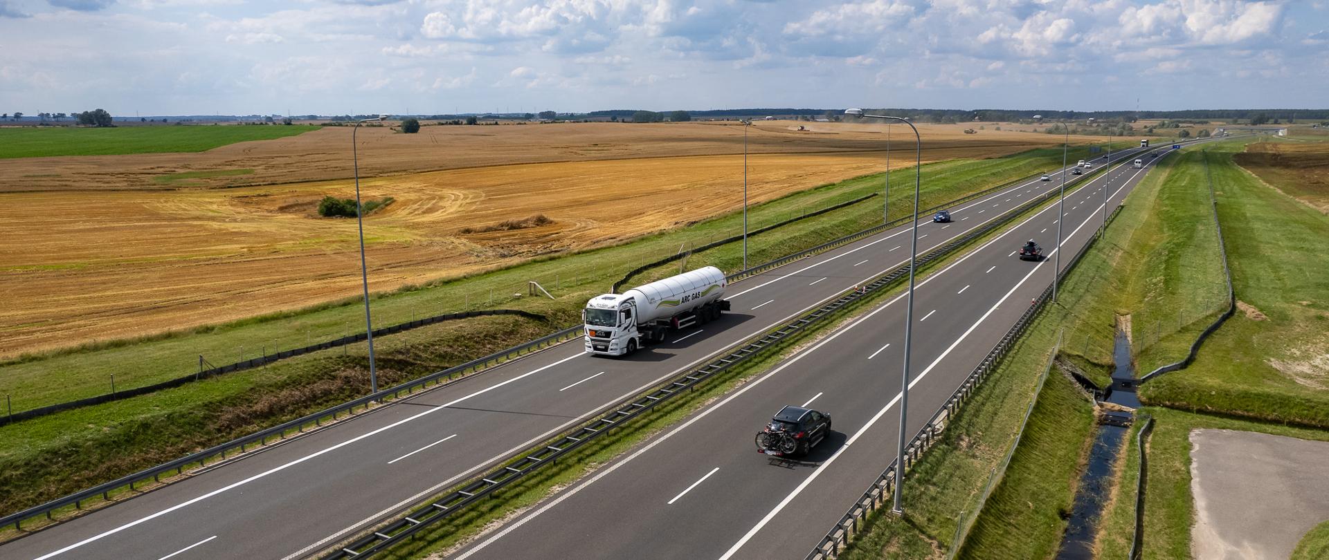 „Lotnicze zdjęcie drogi ekspresowej przebiegającej przez otwarte tereny rolnicze. Na dwujezdniowej trasie z pasem rozdziału poruszają się samochody osobowe oraz ciężarówka-cysterna. Po obu stronach drogi widoczne są pola uprawne, rowy odwadniające i słupy oświetleniowe. Nad krajobrazem rozciąga się błękitne niebo z licznymi białymi chmurami.”