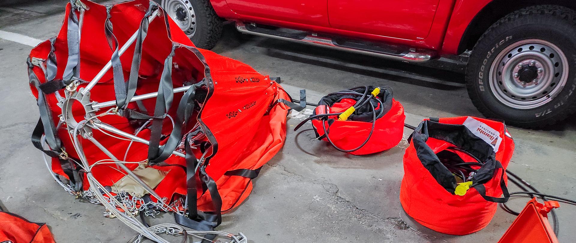 Zdjęcie przedstawia komplet wyposażenia podwieszonego zbiornika pod śmigłowiec typu "Bambi Bucket". Zbiornik koloru pomarańczowego oraz niezbędny osprzęt leży rozłożony na podłodze w garażu jednostki ratowniczo-gaśniczej PSP.