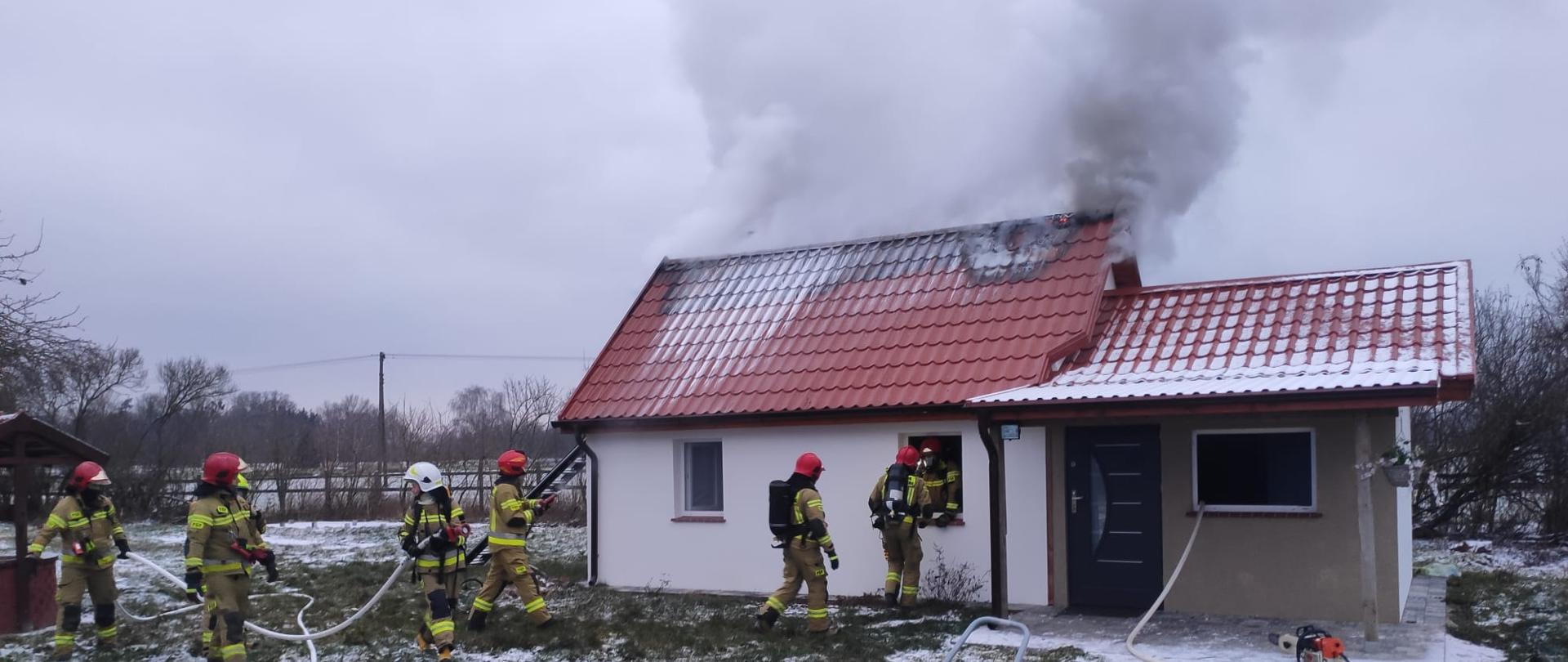 Na zdjęciu budynek który się pali strażacy wchodzący do budynku w aparatach OUO rozwinięte linie gaśnicze i wentylator 