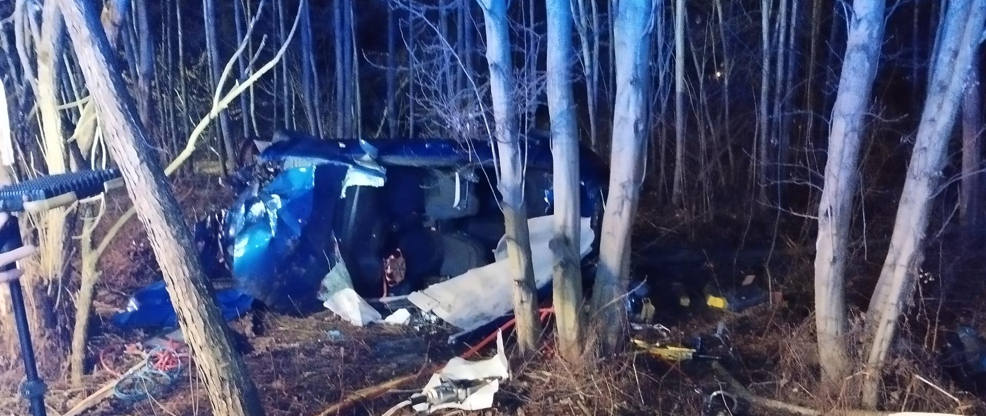 W środku kadru, pośród drzew, znajduje się samochód osobowy leżący na lewym boku (skierowany przodem w lewą stronę) z odciętym i odgiętym do ziemi dachem. Samochód ma zgniecioną do połowy komorę silnika oraz wybite wszystkie szyby. Wokół pojazdu widać rozrzucone elementy karoserii i wyposażenia. Zza lewej strony kadru rozciągnięte są przewody do narzędzi hydraulicznych prowadzące do tylnej części samochodu. Na pierwszym planie w lewym dolnym rogu stoi przenośny maszt oświetleniowy. 