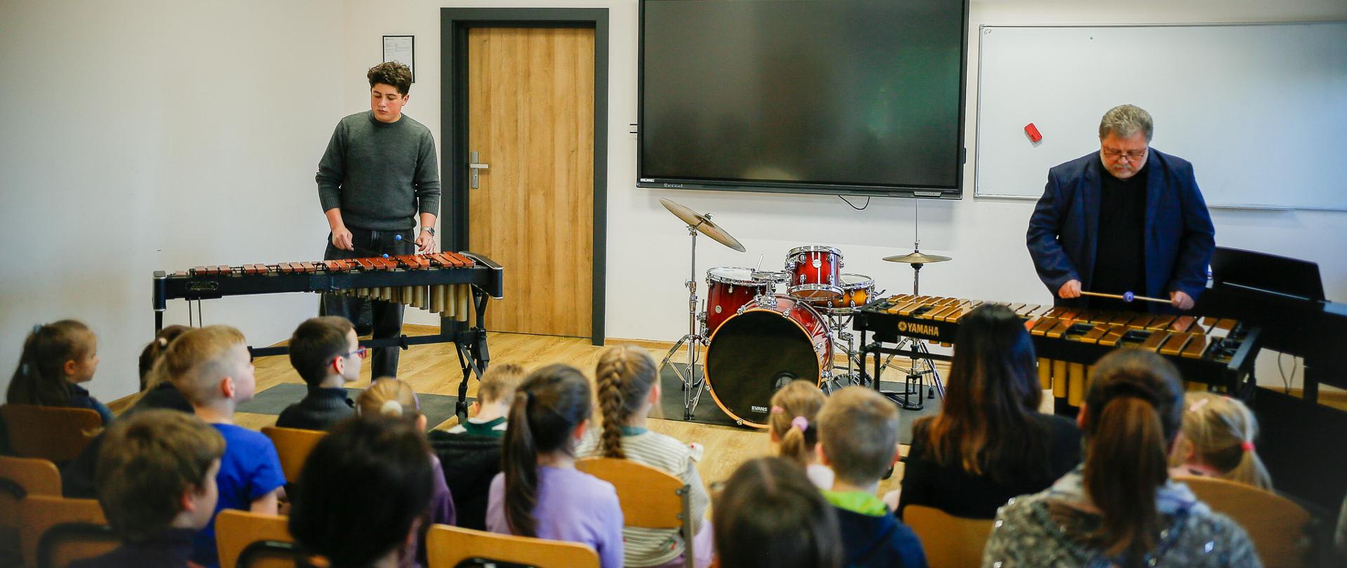 Koncert edukacyjny w szkole. Dwaj mężczyźni prezentują grę na instrumentach sztabkowych (marimba, wibrafon) przed grupą uczniów siedzących w ławkach. W tle widoczna perkusja oraz tablica multimedialna.