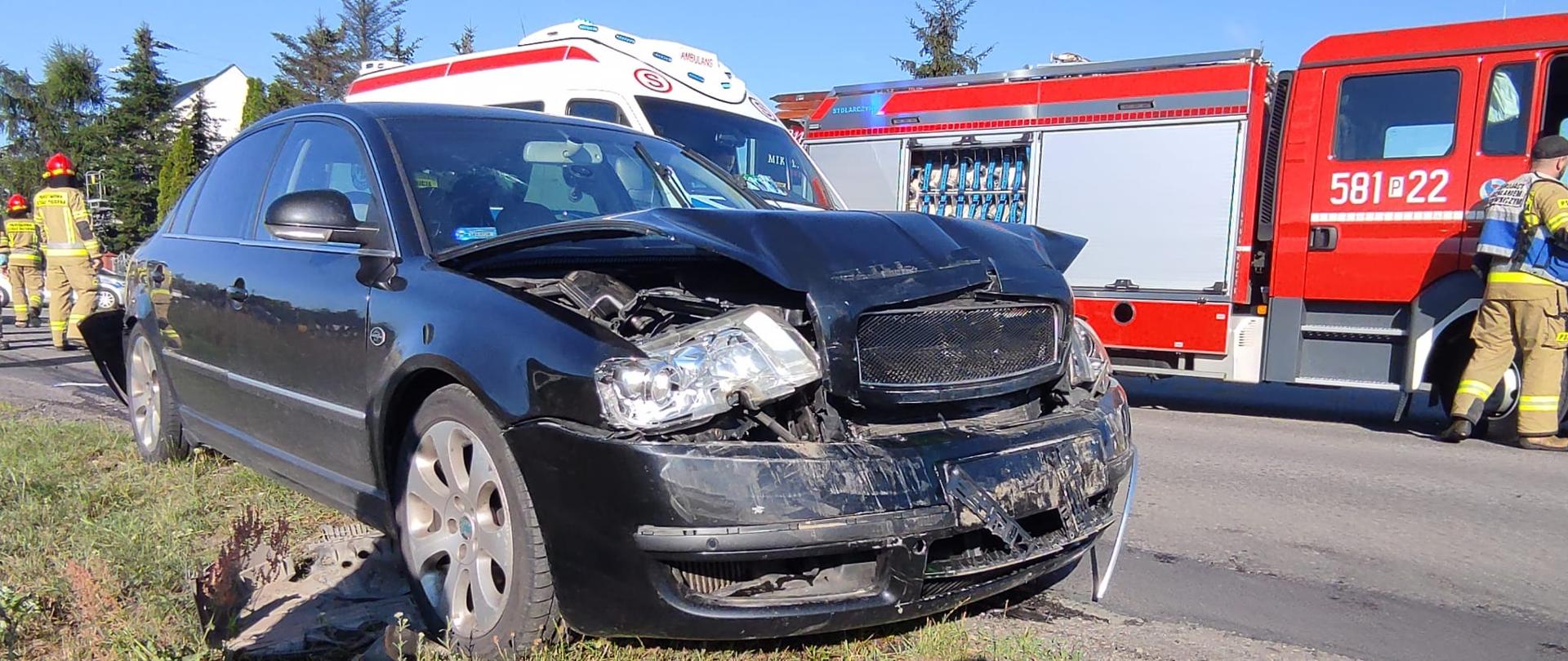 na pierwszym planie czarny samochód osobowy z uszkodzonym przodem za nim widoczny kawałek dachu karetki pogotowie oraz samochód strażacki przy drzwiach strażak za czarnym samochodem inni strażacy