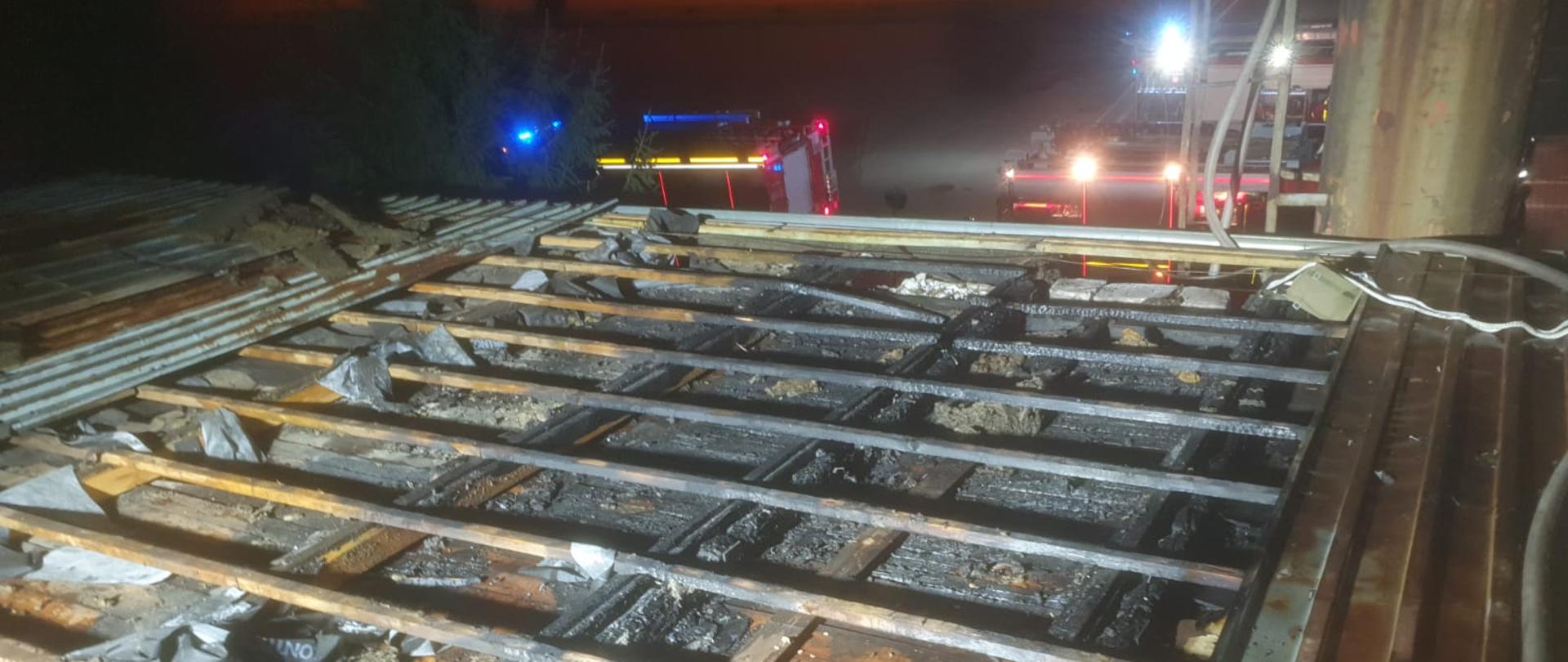 Zdjęcie przedstawia zdemontowaną część pokrycia dachowego z blachy w budynku produkcyjnym oraz spaloną pod nią elementy drewniane. W oddali widoczne są pojazdy pożarnicze