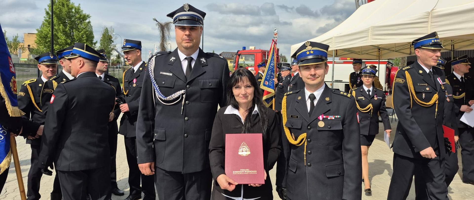 Przedstawiciele OSP i gminy wraz z Komendantem pozują do zdjęcia. W tle strażacy po zakończeniu uroczystego apelu z okazji Dnia Strażaka.