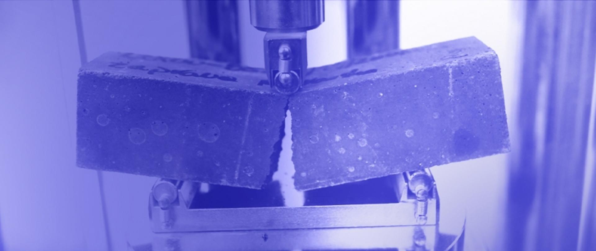 Zdjęcie przedstawia test zginania materiału – blok oparty na dwóch podporach, na który z góry działa siła powodująca jego deformację.