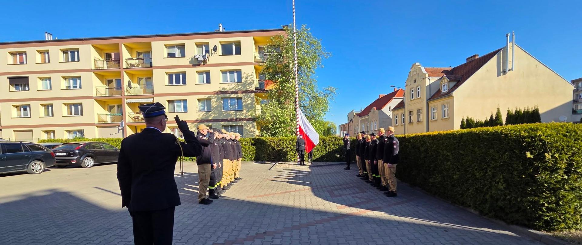Zdjęcie przedstawia strażaków podczas uroczystego podniesienia flagi państwowej na palcu Komendy.
W tle budynki wielorodzinne.
