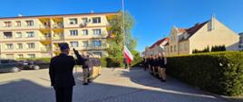 Zdjęcie przedstawia strażaków podczas uroczystego podniesienia flagi państwowej na palcu Komendy.
W tle budynki wielorodzinne.

