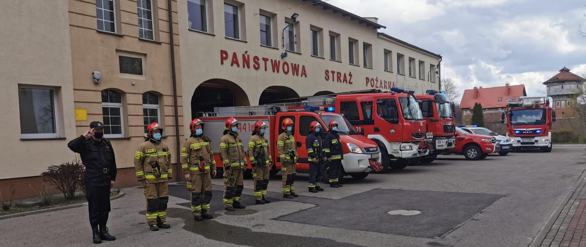 Przed budynkiem komendy stoi na baczność 8 funkcjonariuszy tut. komendy. Za nimi znajdują się wozy strażackie z włączonymi sygnałami świetlnymi.