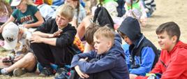 Dzieci siedzące na piaszczystej plaży przyglądające się pokazowi. Dzieci na drugim planie posiadają na szyi zielone chustki. 