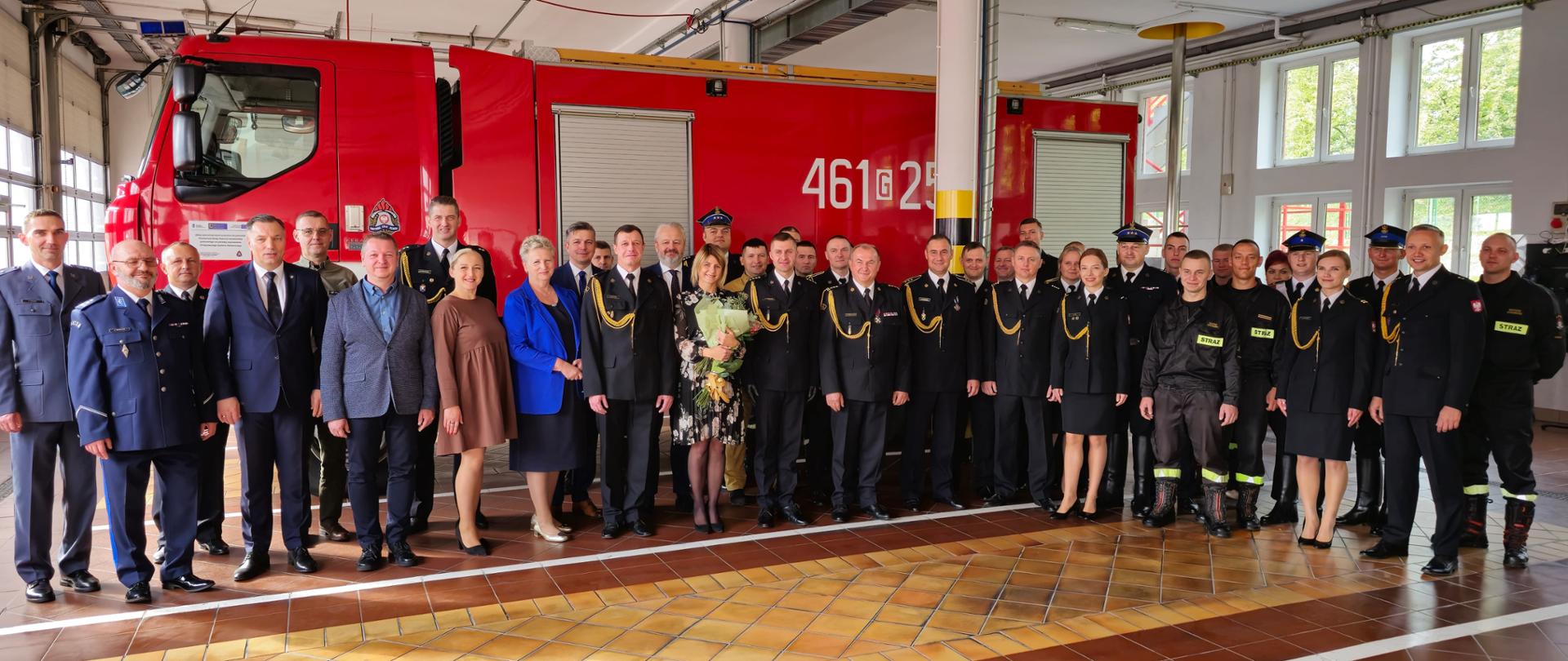 Funkcjonariusze i pracownicy Komendy Powiatowej PSP w Kwidzynie oraz zaproszeni goście pozują do zdjęcia w garażu na tle samochodu pożarniczego.
