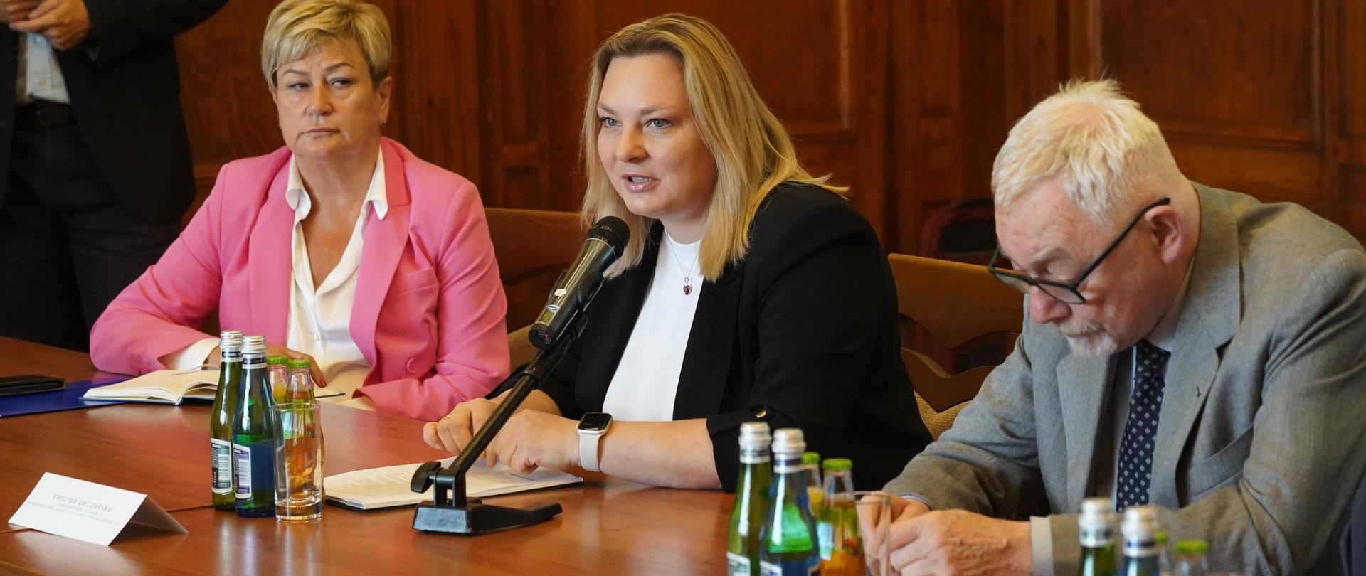 Trzy osoby siedzą przy stole. W środku siedzi i mówi do mikrofonu wiceminister Ewelina Owczarska. 