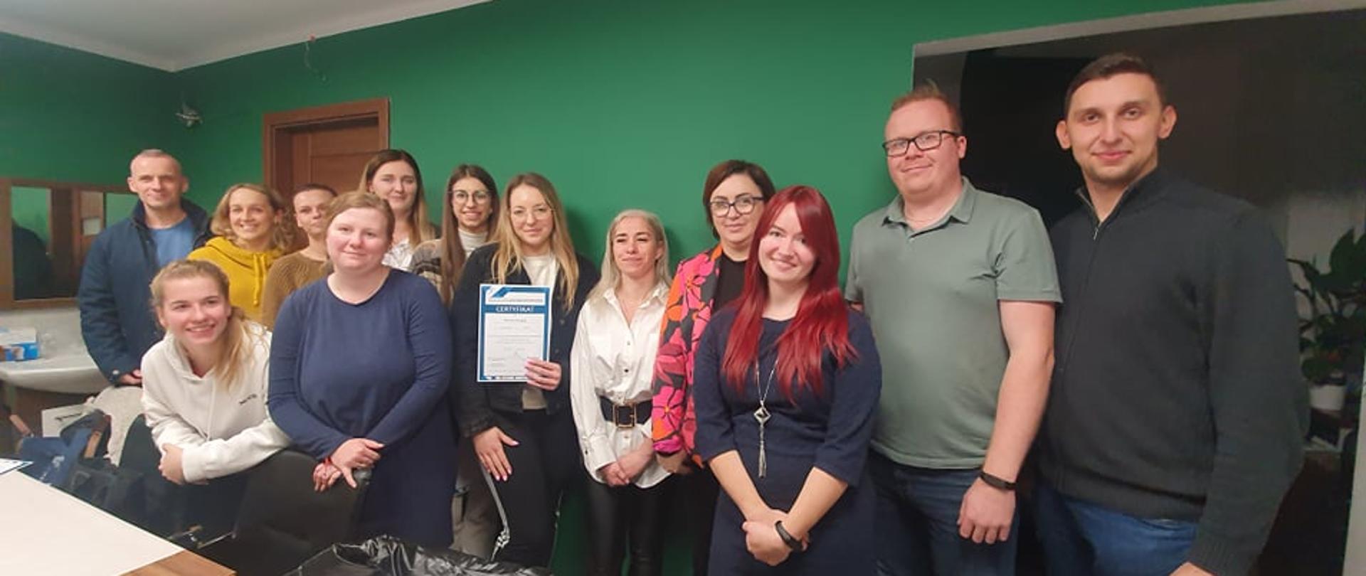 Zdjęcie przedstawia grupę kilkunastu osób, które znajdują się w pokoju, gdzie zakończył się egzamin z języka migowego. Wszyscy uśmiechają się do zdjęcia. W tle zielona ściana pokoju.