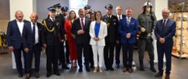 Zdjęcie grupowe, na którym widoczni są zaproszeni goście. Fotografię wykonano w budynku remizy OSP Pawłosiów, w której znajduje się sala pamięci. Goście ubrani są w stroje wyjściowe i wizytowe. Widoczne są również manekiny ubrane w umundurowanie strażackie z dawnych lat.