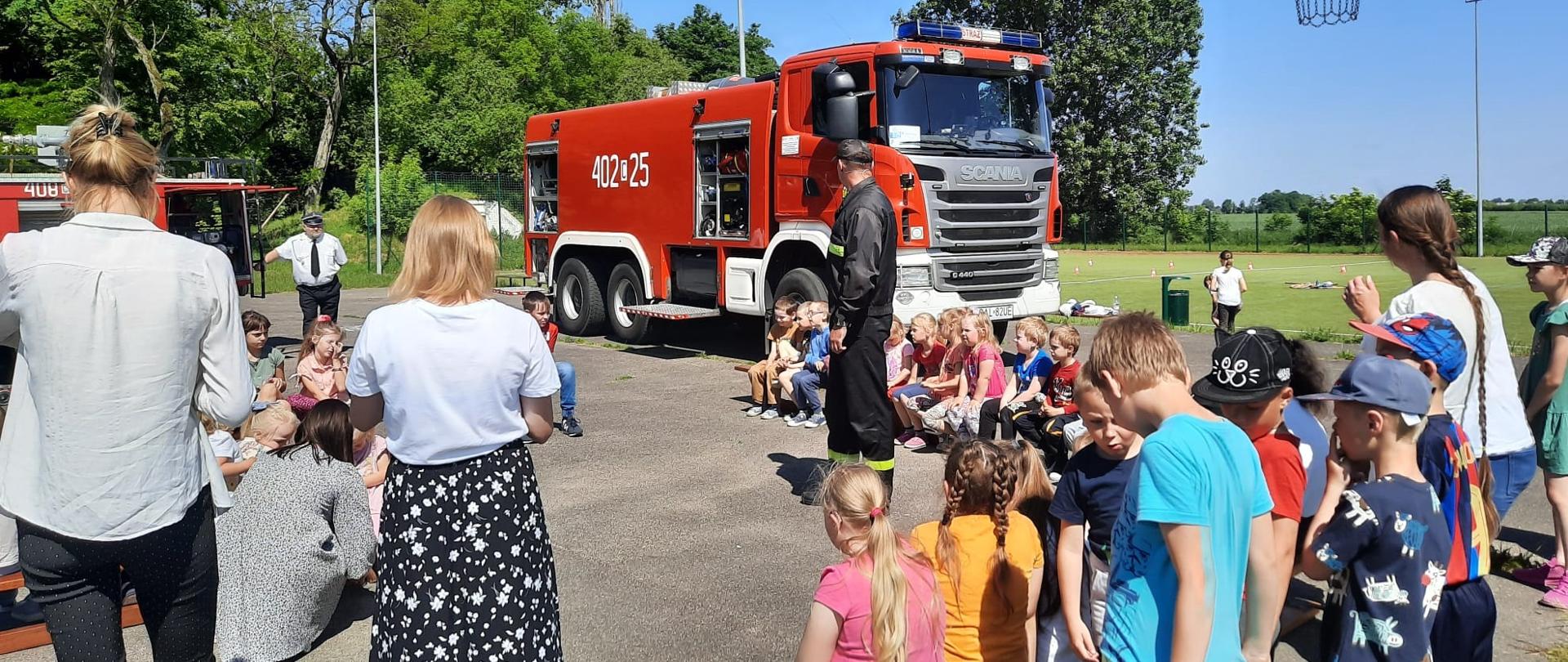 Na zdjęciu widać grupę uczniów na płycie boiska, którzy siedzą na ławkach w okręgu. Wśród nich stoi strażak, który prowadzi pogadankę. W tle widać samochód pożarniczy.