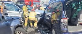 Na zdjęciu znajdują się wraki pojazdów oraz strażacy działający przy pojazdach, w tle widać samochód strażacki oraz inne osoby