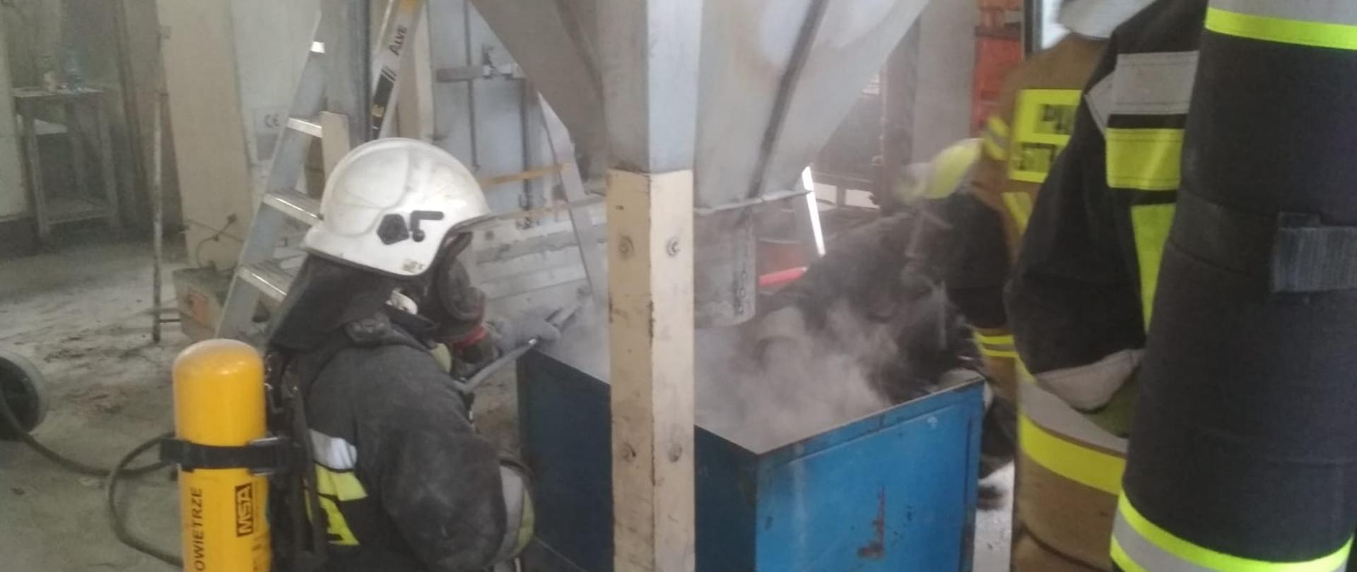 Na zdjęciu widoczni strażacy w ubraniach specjalnych i hełmach na głowach z założonymi aparatami ochrony dróg oddechowych na plecach podczas pożaru. Pożar miał miejsce w hali zakładu pracy w Praszce. W centralnej części kadru widoczna część instalacji technologicznej po pożarze. Na drugim planie widoczna rozstawiona drabina aluminiowa.