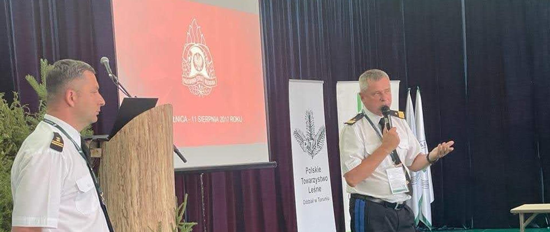 Pomorski komendant wojewódzki Państwowej Straży Pożarnej w obecności strażaka stojącego obok trzymając mikrofon przemawia. Za nim na ekranie logo Państwowej Straży Pożarnej.