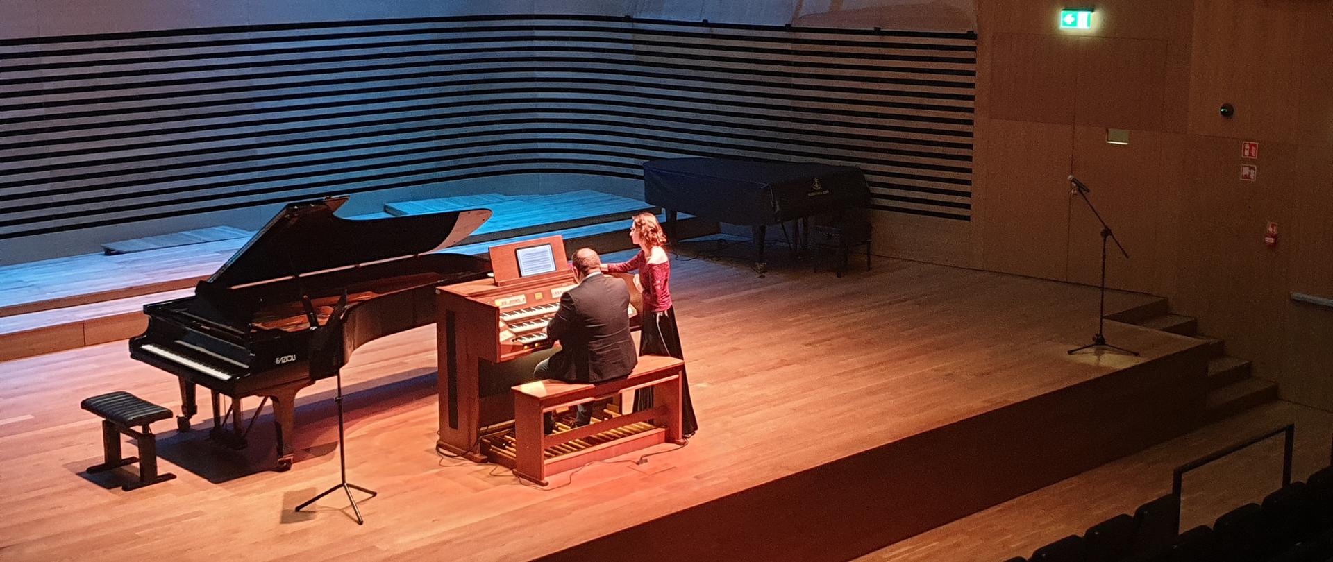 Mężczyzna siedząc tyłem do publiczności gra na organach, kobieta stoi obok na scenie sali koncertowej PSM