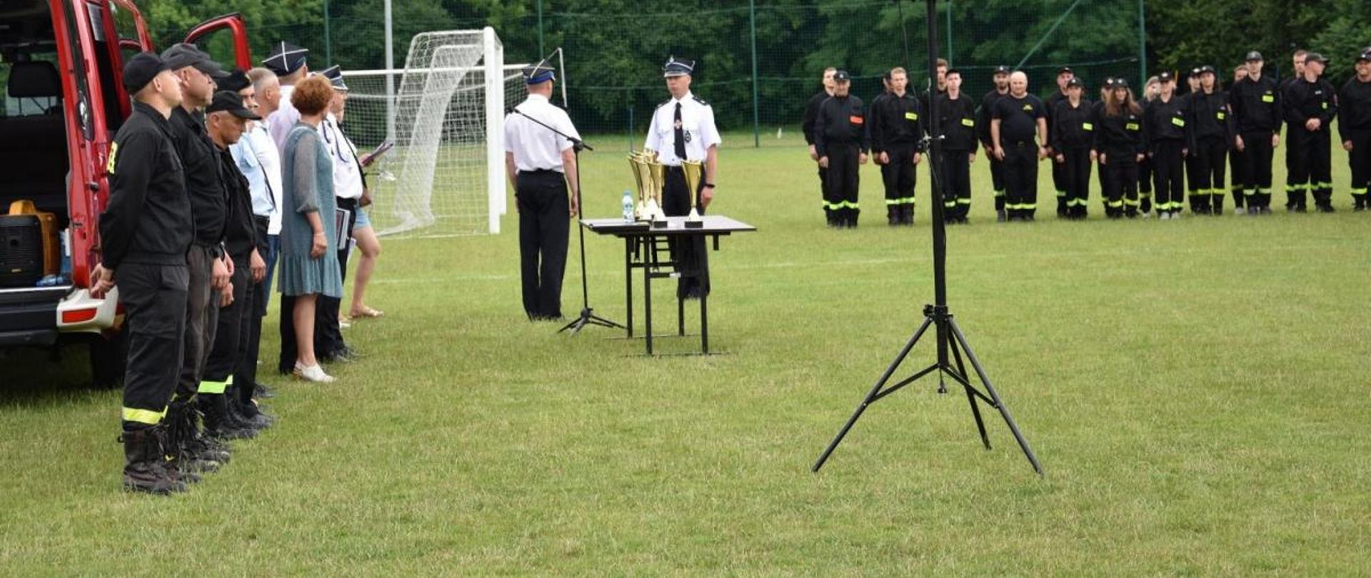 Międzygminne Zawody Sportowo – Pożarnicze jednostek OSP z terenu gmin Sokołów Podlaski i Bielany i miasta Sokołowa Podlaskiego. na zdjęciu strażacy w ubraniu koszarowym stoją w szeregu na zbiórce na boisku trawiastym. na rodu stoi statyw z głośnikiem i stół z pucharami.