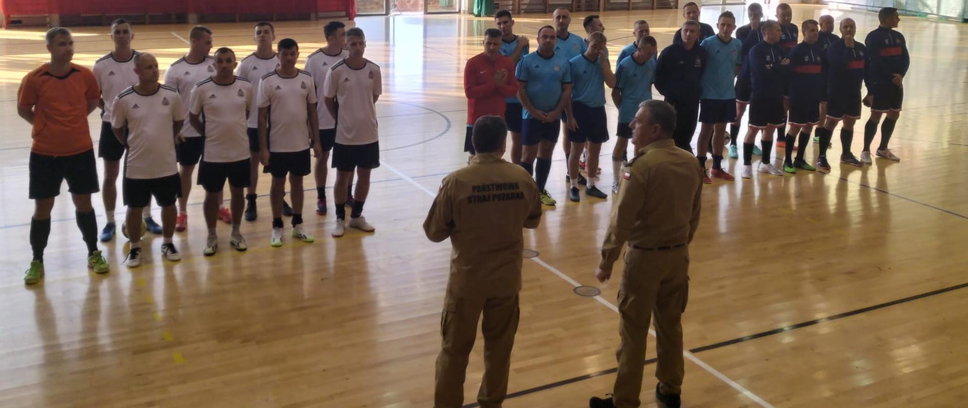 Na 1 planie stoją tyłem komendanci ubrani w ubrania koszarowe koloru piaskowego, w tle w strażacy biorący udział w turnieju halowej piłki nożnej ubrani są w stroje sportowe. Znajdują sie na parkiecie hali sportowej, w tle okna. 