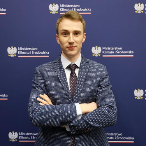 Na zdjęciu znajduje się młody mężczyzna na niebieskim tle z logo Ministerstwa Klimatu i Środowiska. Ubrany jest w białą koszulę oraz granatową marynarkę z białymi wzorami. Na szyi zawiązany jest brązowy krawat w białe wzory.