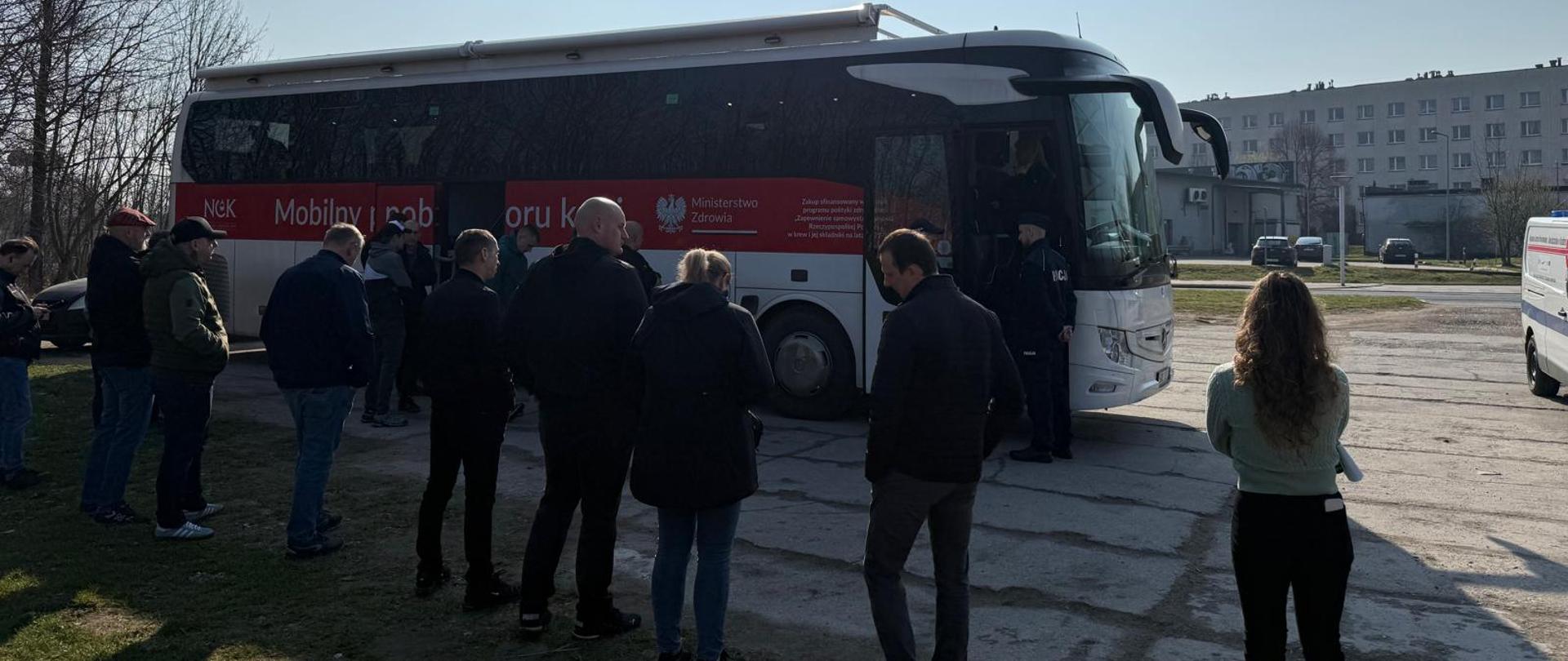 Widoczni ludzie stojący luźno przed autobusem do poboru krwi. Za autobusem w tle widoczne bloki mieszkalne i drzewa.