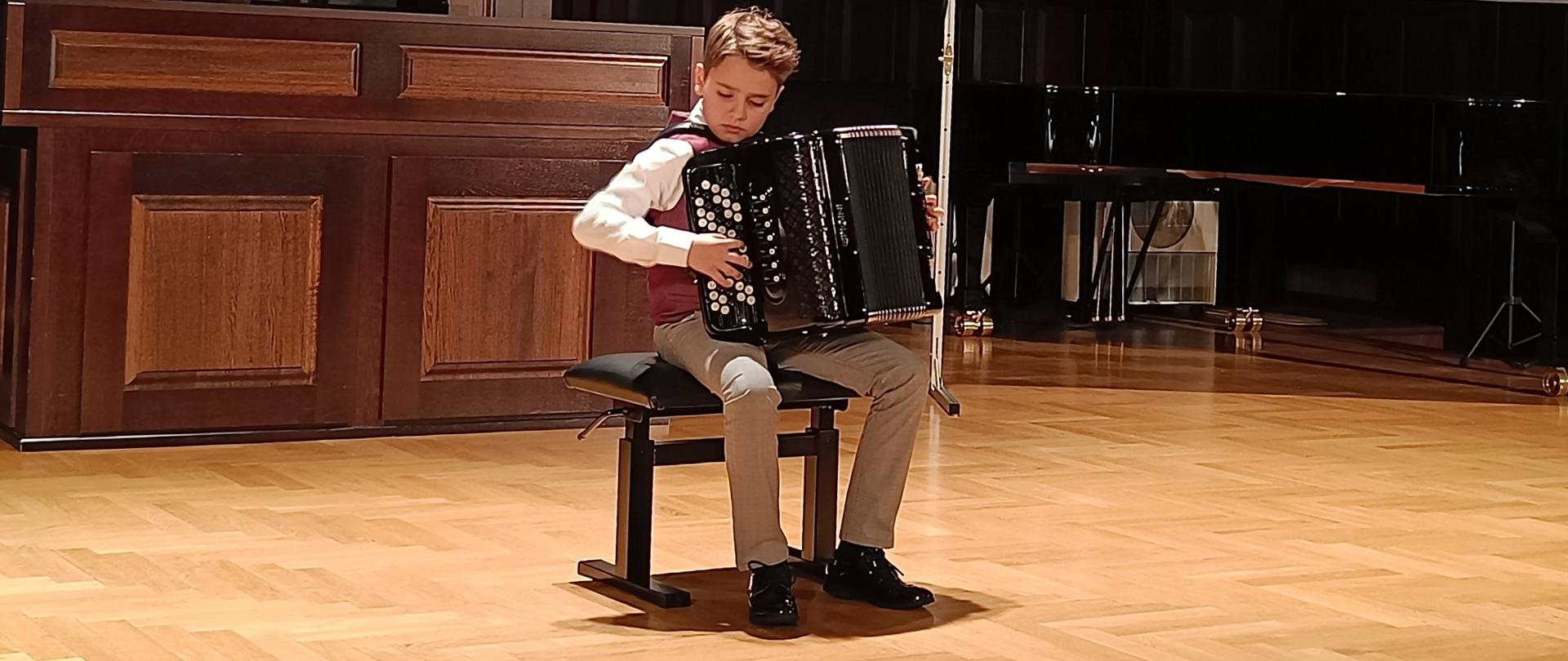 Uczeń siedzący na czarnym krześle, grający na akordeonie. Z tyłu widoczne organy w kolorze brązowym. Po prawej stronie rzutnik z wyświetlonym slajdem: 40 lat katedry akordeonu 1983-2023, Młoda Katedra Akordeonu, Akademia Muzyczna im. Karola Szymanowskiego w Katowicach.
