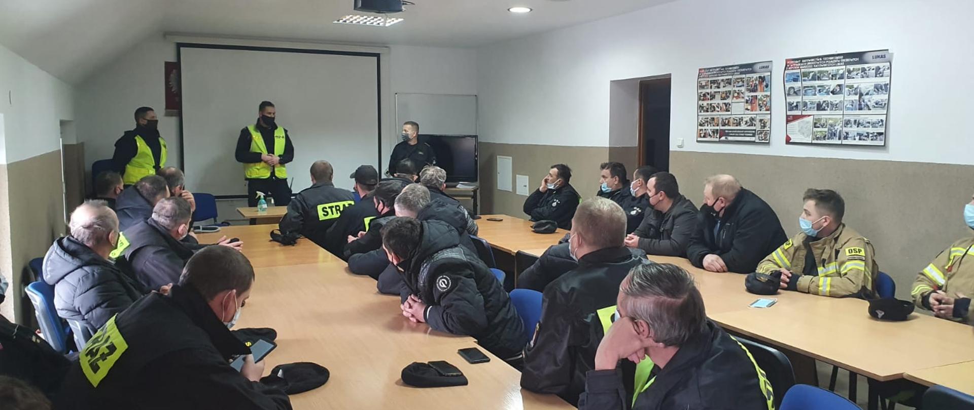 Zdjęcie przedstawia pomieszczenie Komendy Powiatowej Państwowej Straży Pożarnej, strażaków Ochotniczych Straży Pożarnych, funkcjonariuszy policji oraz Państwowej Straży Pożarnej podczas szkolenia kierowców pojazdów uprzywilejowanych.
