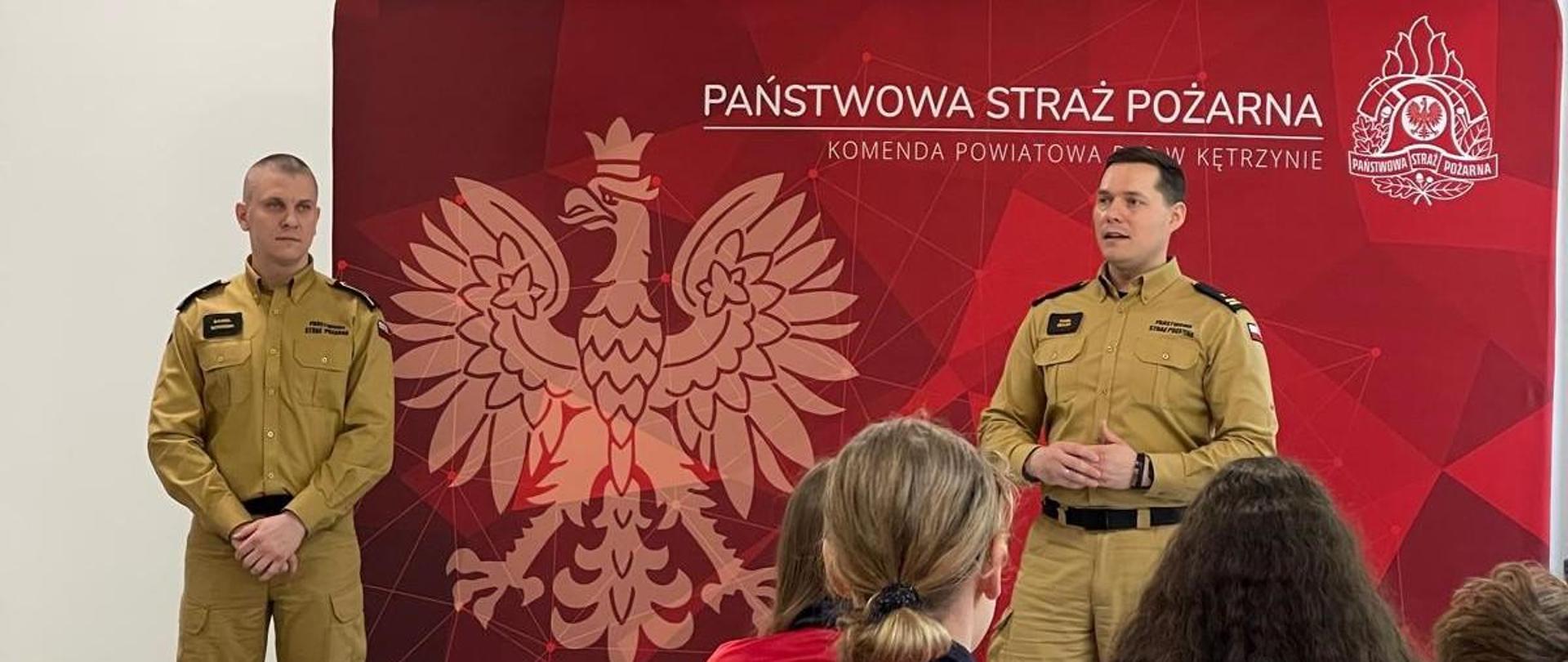 Na zdjęciu widać dwóch strażaków na tle czerwonego banneru. Tyłem siedzą dwie osoby w bluzie z napisem Młodzieżowa Drużyna Pożarnicza.