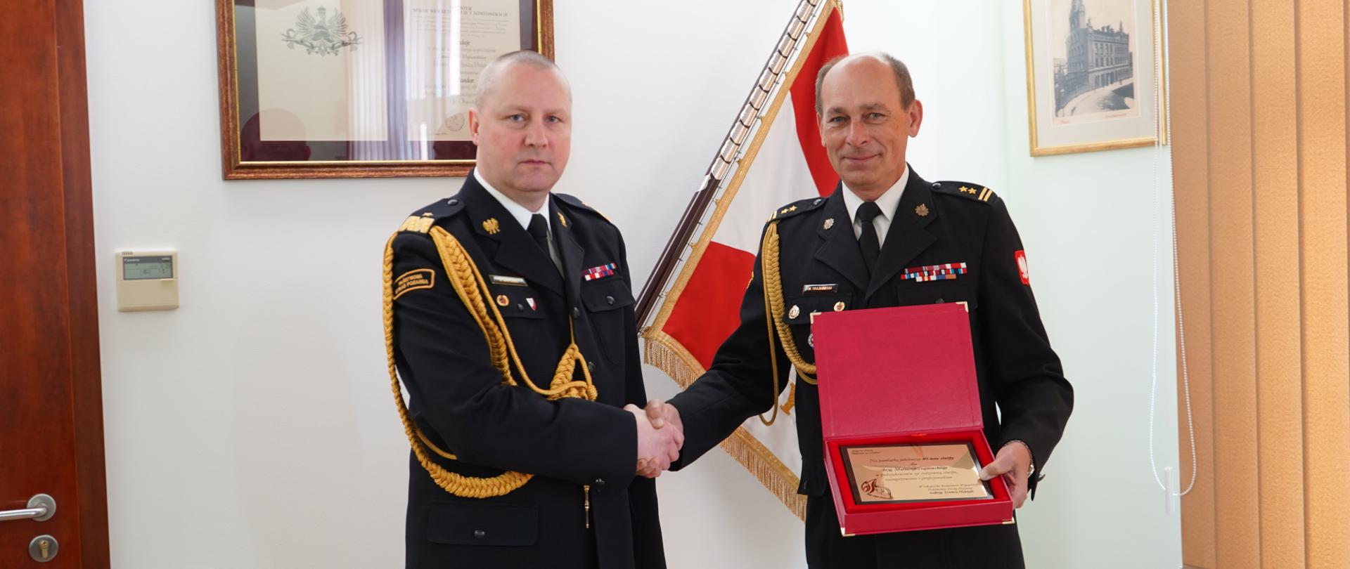 dwóch strażaków ściska sobie dłoń, jeden z nich trzyma w ręku grawer, w tle sztandar