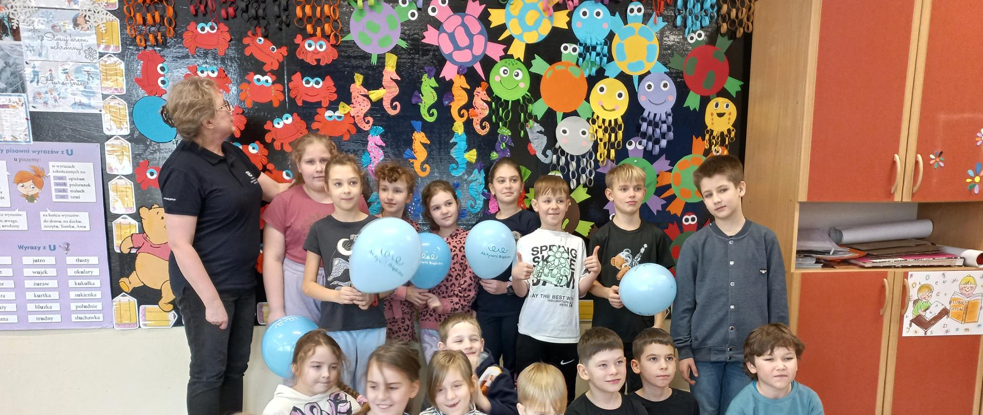 Klasa. Uczniowie pozują do zdjęcia z pracownikiem Wód Polskich. W tle tablica z pracami plastycznymi.