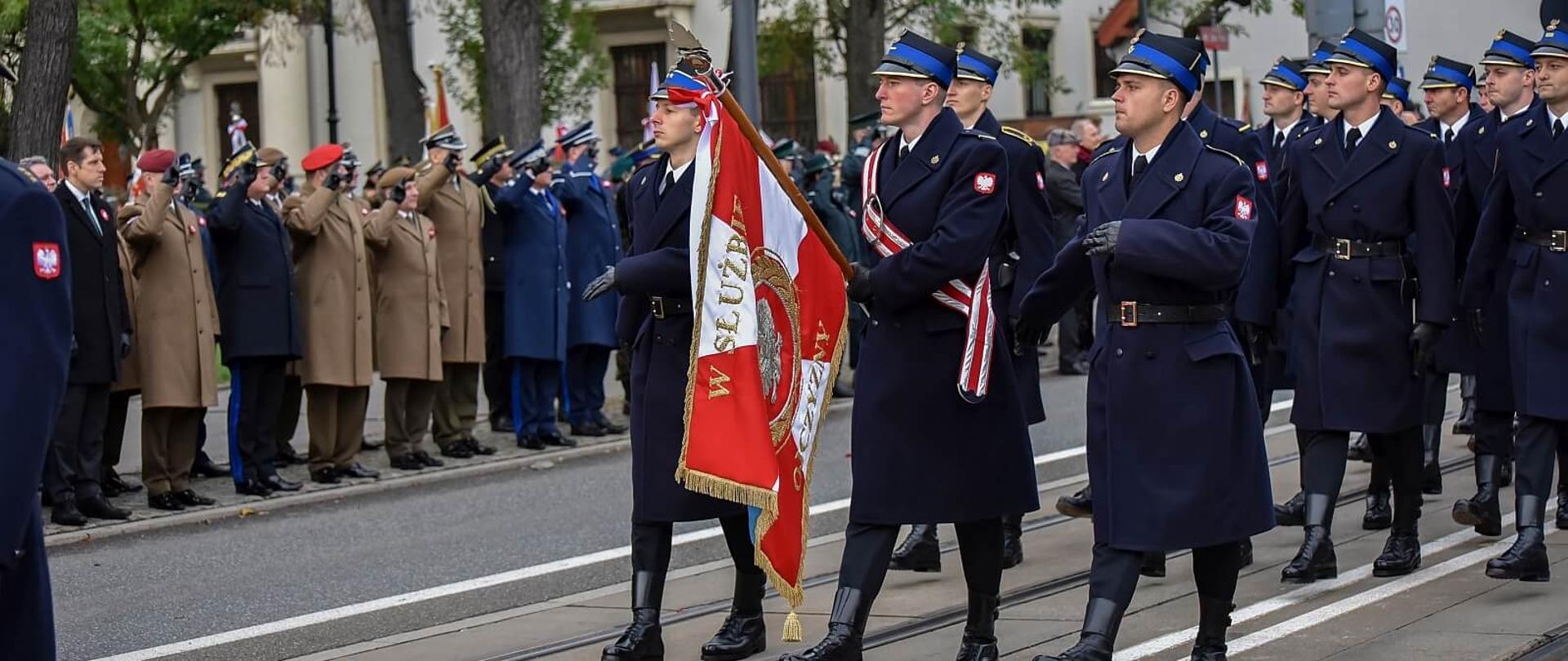 poczet i kompania honorowa komendy wojewódzkiej PSP w Łodzi maszerują w trakcie defilady na drugim planie komendanci różnych służb odbierają defiladę