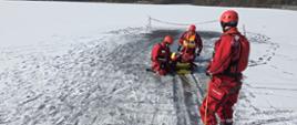 Czterech ratowników w czerwonych kombinezonach i kaskach prowadzi ćwiczenia na zamarzniętym akwenie. dwóch klęczy przy osobie leżącej na żółtej desce ratowniczej w pobliżu przerębla, a trzeci stoi z boku i trzyma linę asekuracyjną; teren wokół jest ośnieżony, w tle widać drzewa i taśmę zabezpieczającą miejsce działań.