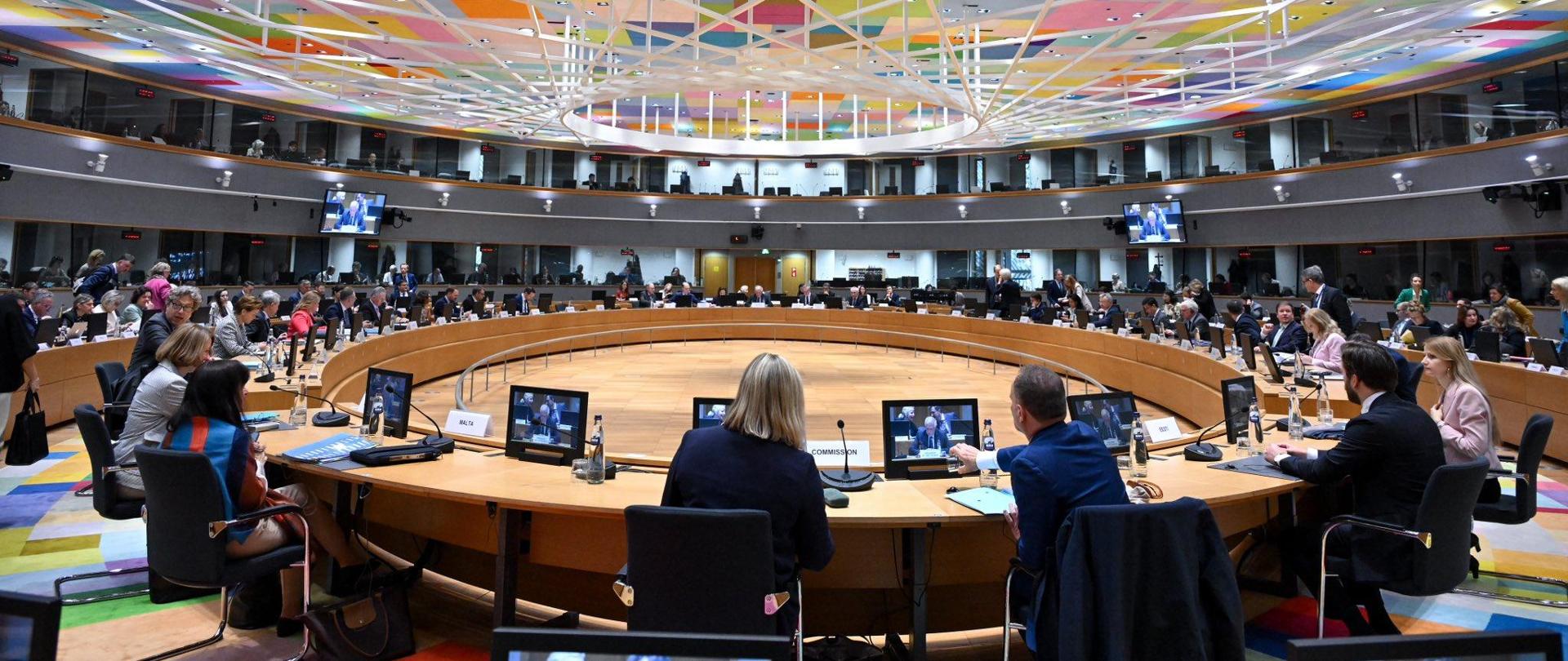 Sala posiedzeń z okrągłym stołem przy którym siedzą delegaci, każdy z monitorem przed sobą.