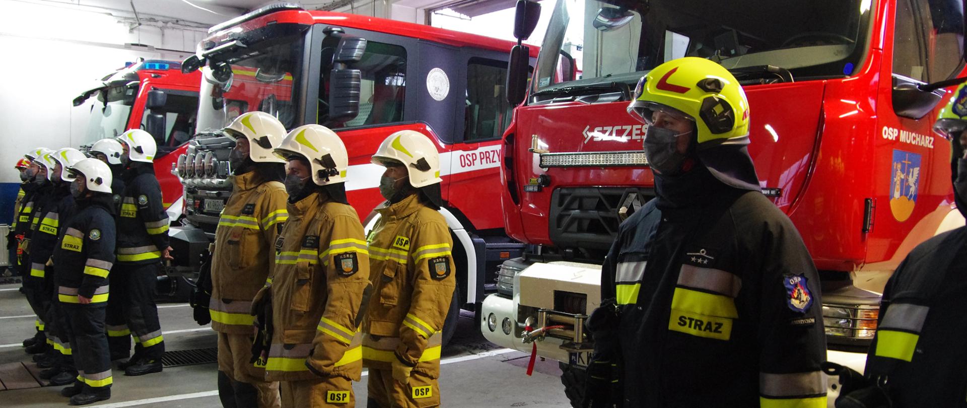 Widok z przodu. Strażacy PSP i OSP w ubraniach specjalnych stoją w przed pojazdami ratowniczo-gaśniczymi.