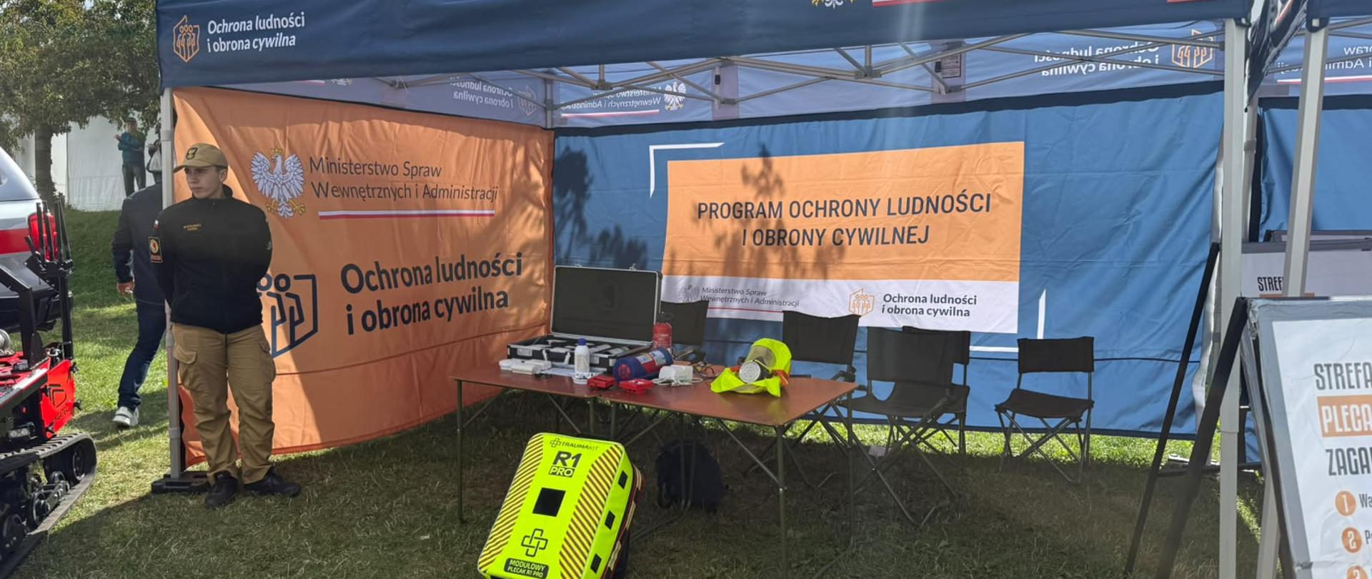 Granatowo-pomarańczowy namiot z logom i napisem Ochrona ludności i obrona cywilna oraz MSWiA pod którym stoi stolik z leżącymi przedmiotami ochronnymi, przed namiotem wystawiony jest modułowy plecak R1Pro, po lewej stronie stojący kadet CS PSP