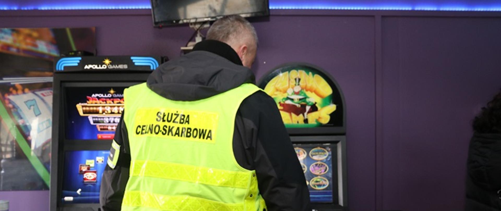 Funkcjonariusz po cywilnemu, w żółtej kamizelce z napisem Służba Celno-Skarbowa stoi przed automatami hazardowymi.