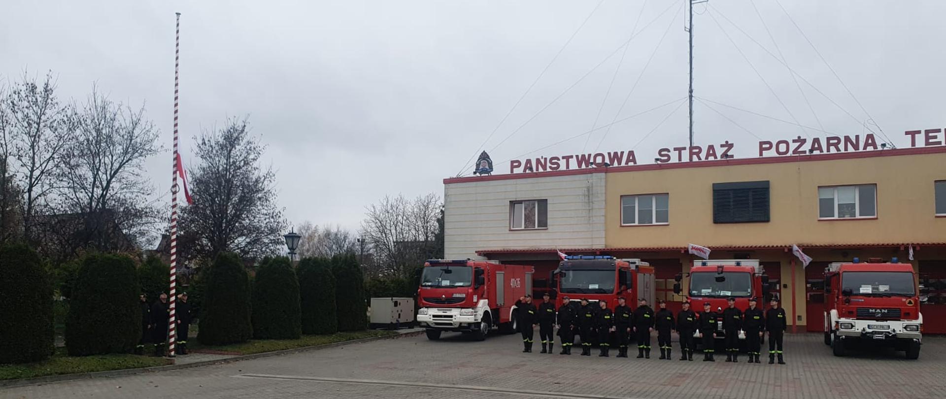 Plac manewrowy pokryty kostką brukową. W tle budynek komendy powiatowej w kolorze żółtym z napisem na dachu Państwowa Straż Pożarna. Przed budynkiem wyprowadzone samochody strażackie koloru czekowego. Strażacy stoją w szeregu, dowódca oddaje honory. W lewej strony kadru widoczny maszt flagowy a nic wciągana go góry flaga