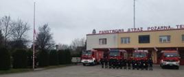 Plac manewrowy pokryty kostką brukową. W tle budynek komendy powiatowej w kolorze żółtym z napisem na dachu Państwowa Straż Pożarna. Przed budynkiem wyprowadzone samochody strażackie koloru czekowego. Strażacy stoją w szeregu, dowódca oddaje honory. W lewej strony kadru widoczny maszt flagowy a nic wciągana go góry flaga