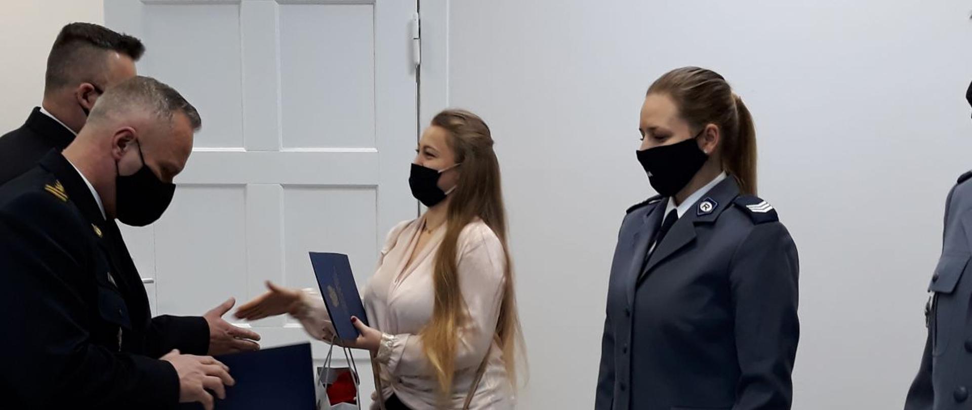 Na zdjęciu widoczne 4 osoby w trakcie wręczania teczek z podziękowaniami i upominków w jasnym pomieszczeniu. Po lewej dwaj strażacy w galowych czarnych mundurach po prawej 2 kobiety. Od lewej kobieta o długich jasnych rozpuszczonych włosach, w jasnej bluzce i ciemnych spodniach. Po prawej policjantka z kitką na głowie w mundurze galowym: niebieska marynarka i czarna spódnica. Wszystkie osoby na twarzy mają założone czarne maseczki. W tle widoczne białe drzwi do pomieszczenia.