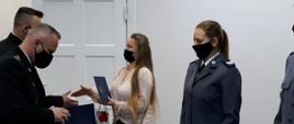 Na zdjęciu widoczne 4 osoby w trakcie wręczania teczek z podziękowaniami i upominków w jasnym pomieszczeniu. Po lewej dwaj strażacy w galowych czarnych mundurach po prawej 2 kobiety. Od lewej kobieta o długich jasnych rozpuszczonych włosach, w jasnej bluzce. Po prawej policjantka z kitką na głowie w mundurze galowym: -niebieska marynarka. Wszystkie osoby na twarzy mają założone czarne maseczki. W tle widoczne białe drzwi do pomieszczenia.