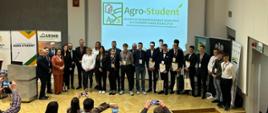 Zdjęcie przedstawia dużą grupę osób pozujących na scenie w auli wykładowej. Grupa składa się z uczniów w garniturach trzymających dyplomy oraz dorosłych organizatorów. Nad nimi, na dużym ekranie projekcyjnym, wyświetlane jest logo „Agro-Student” z podpisem „XII EDYCJA OGÓLNOPOLSKIEGO KONKURSU DLA UCZNIÓW SZKÓŁ ROLNICZYCH”. Na scenie po lewej stronie widać również banner z logo ARiMR. W pierwszym planie widoczne są sylwetki widzów robiących zdjęcia telefonami.