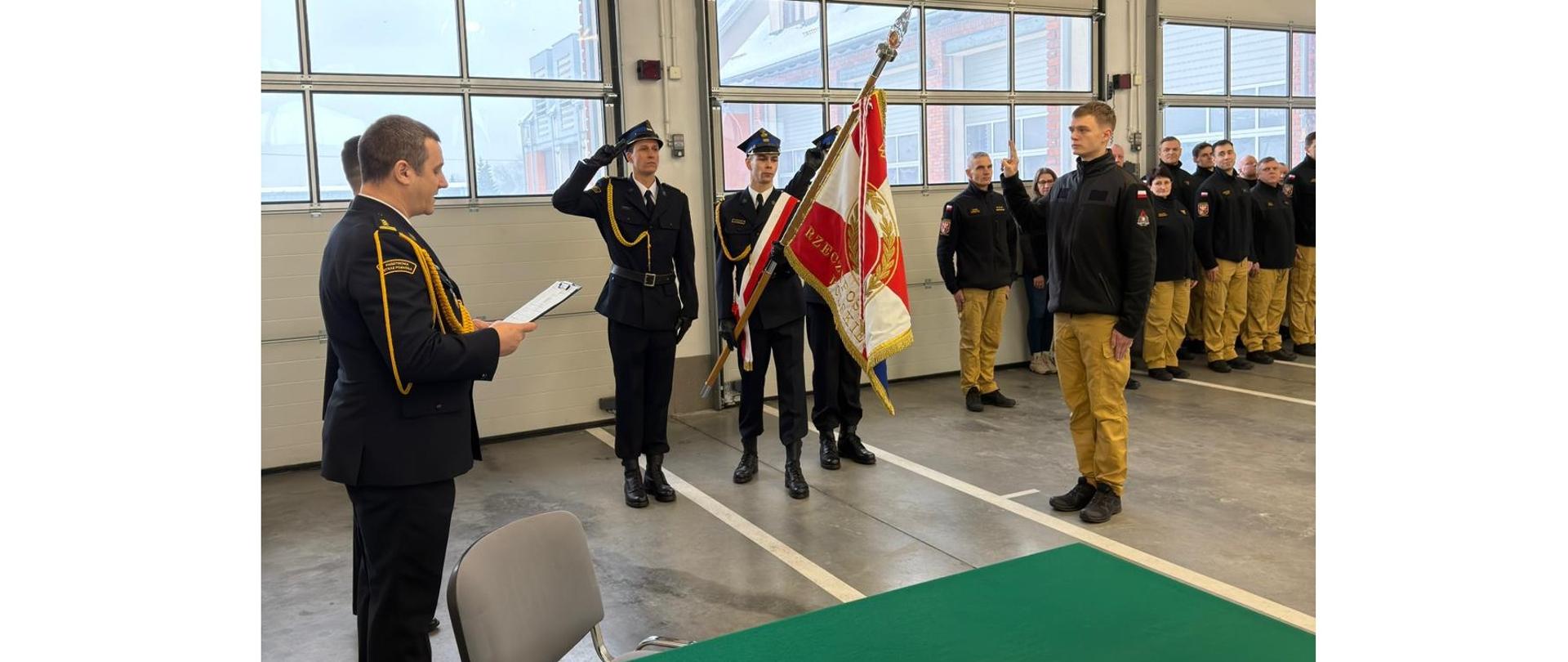 Zdjęcie przedstawia uroczysty moment składania ślubowania przez nowo przyjętego strażaka Wojciecha Wiśniewskiego do służby w Komendzie Powiatowej Państwowej Straży Pożarnej w Gołdapi oraz sztandar komendy. W tle można zaobserwować pozostałych strażaków biorących udział w uroczystej zbiórce